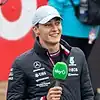 George Russell wearing a silver baseball cap on his ead, a black coat with sponsors logos and he is holding a microphone in his right hand