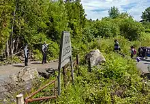 On the left side of a sign, which slightly side says "Road Closed" in the foreground, one of two police officers points to his left. On the right side, two people have wheeled suitcases behind them.