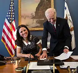 Julia Louis-Dreyfus seated at a desk, with Biden standing over her, both laughing.
