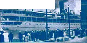 Blue-tinted photograph of a ship, with many people standing in the foreground.