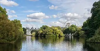 Photograph looking east over St. James's Park Lake from the Blue Bridge, with the London Eye and parks of White Hall visible above the trees in the distance.